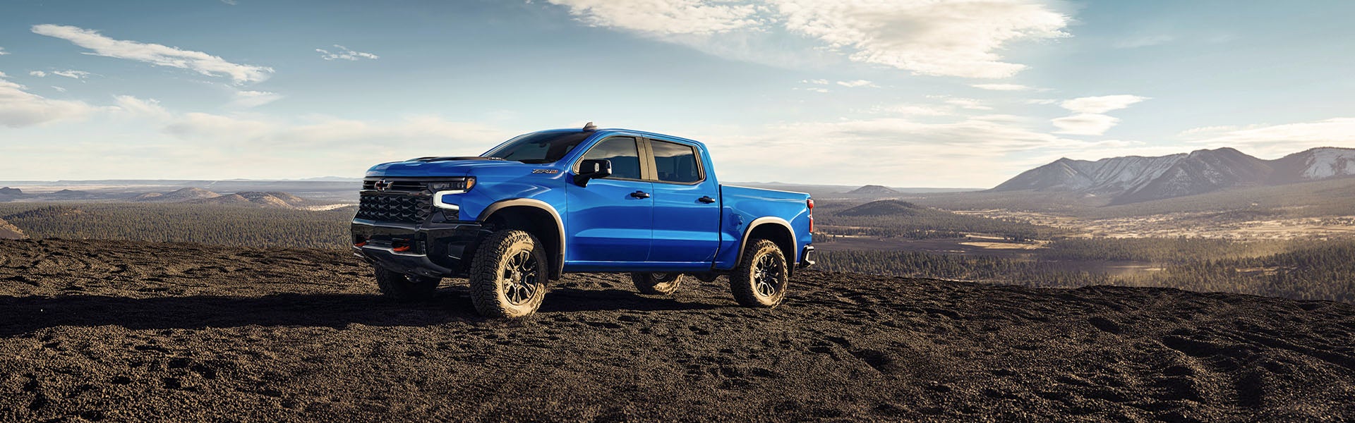 A blue Chevrolet Silverado 1500 parked in front of a scenic mountain landscape with a clear sky in the background.